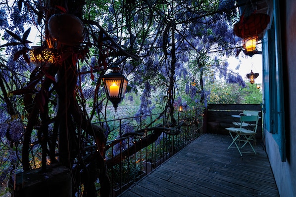A wooden deck with a railing, a table, and chairs, surrounded by trees and purple flowers.