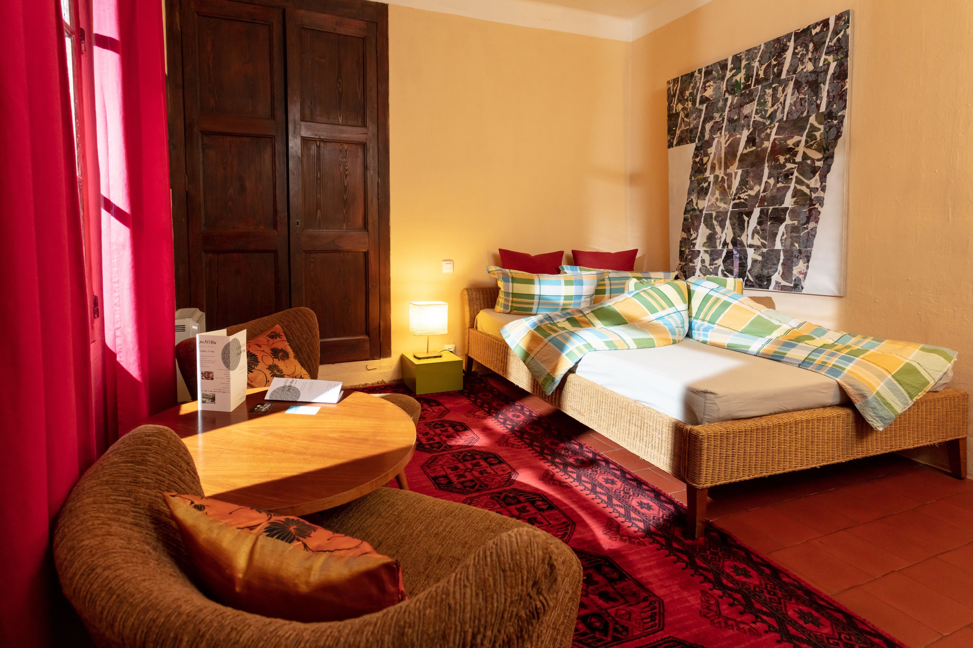 A hotel room with a bed, a chair, a table, a lamp, and a large tapestry on the wall.
