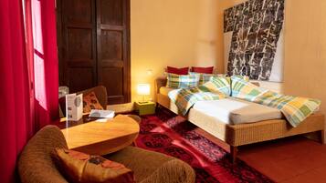 A hotel room with a bed, a chair, a table, a lamp, and a large tapestry on the wall.
