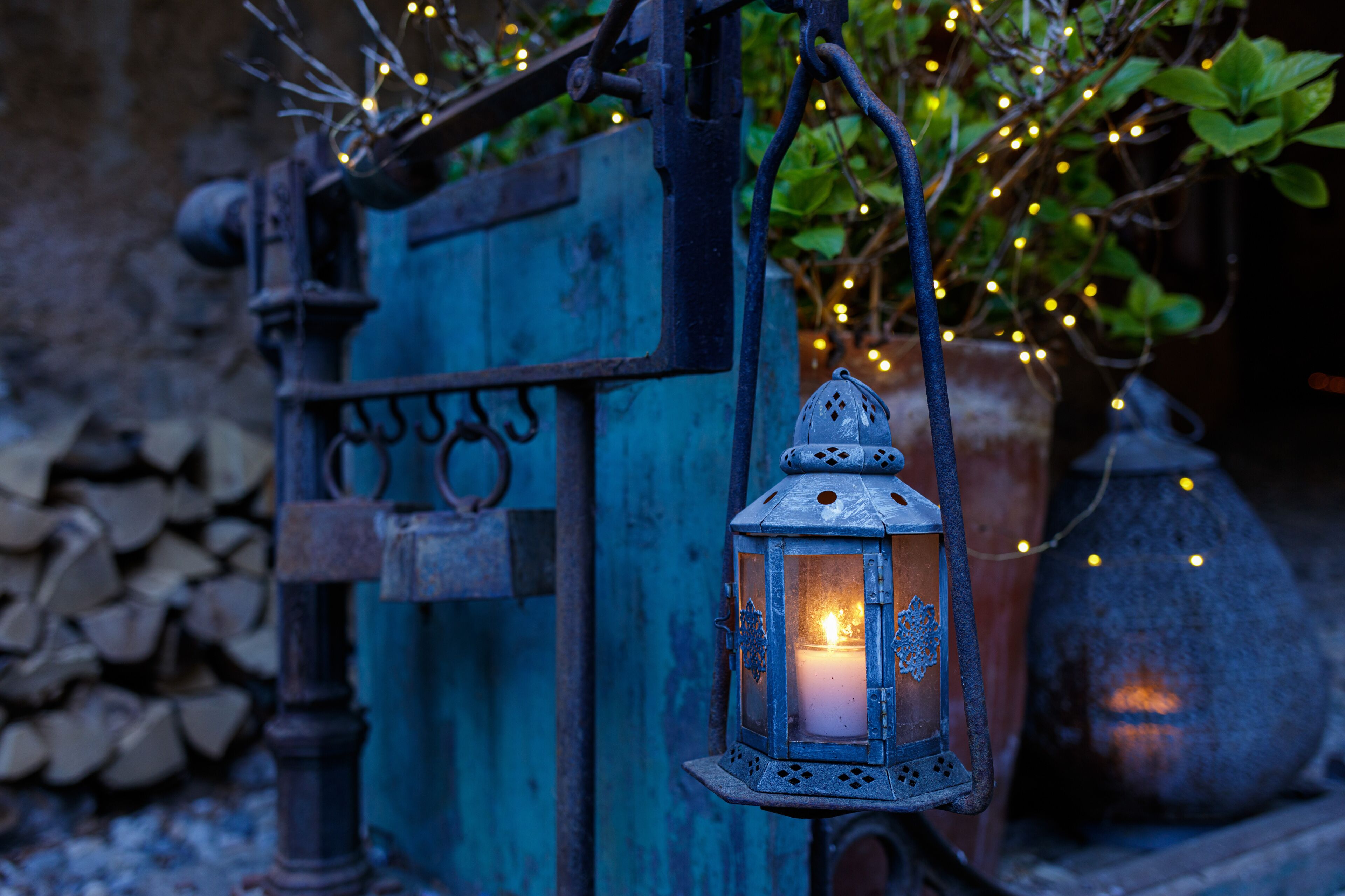 A rustic lantern with a lit candle, hanging from a metal structure with a blue door and a potted plant in the background.