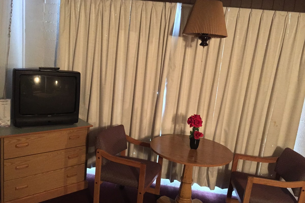 A room with a wooden TV stand, a small table with a vase of flowers, and two chairs.