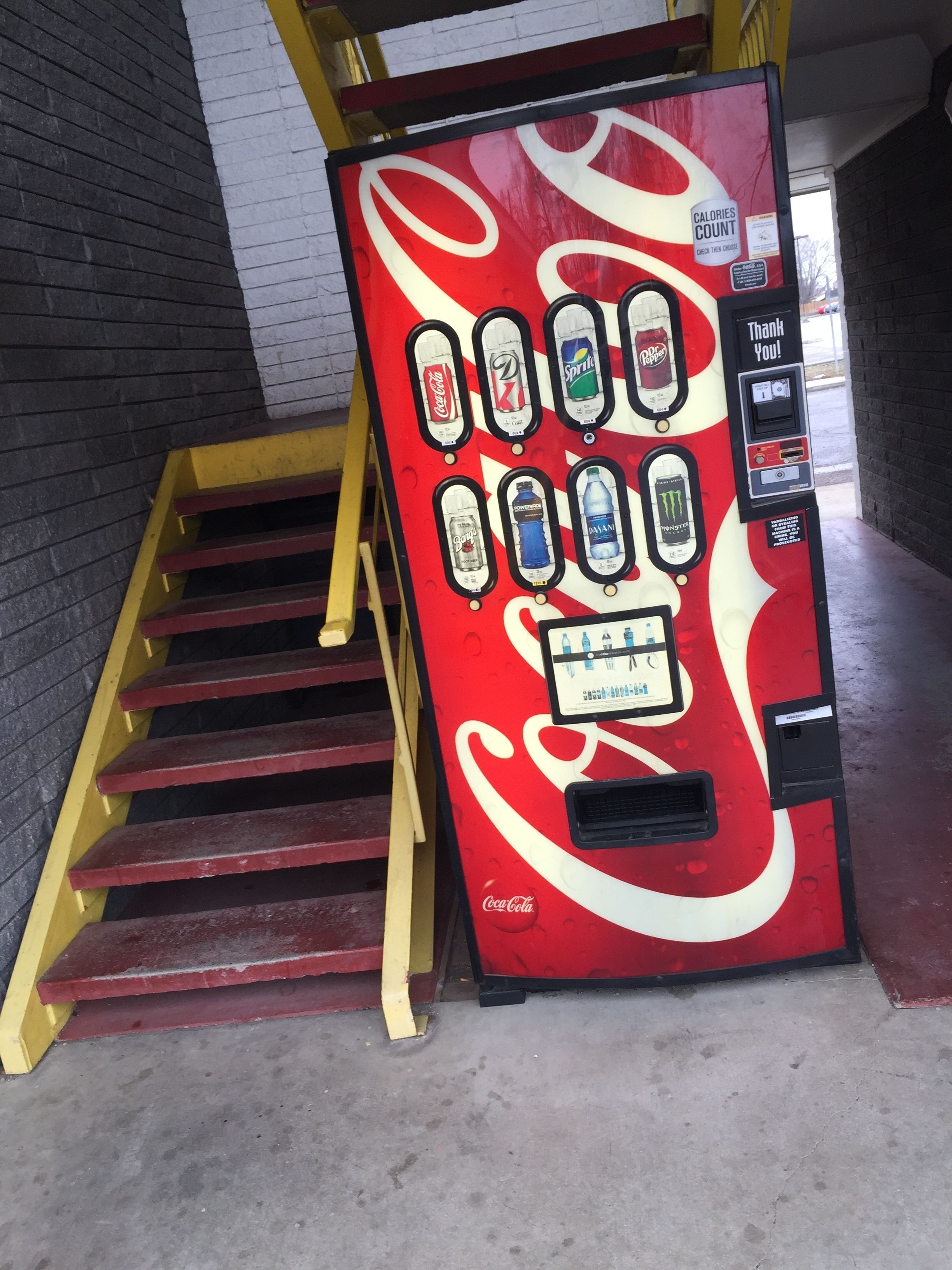 A Coca-Cola branded vending machine with a smiley face design, located next to a staircase.