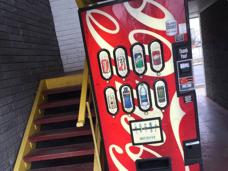 A Coca-Cola branded vending machine with a smiley face design, located next to a staircase.