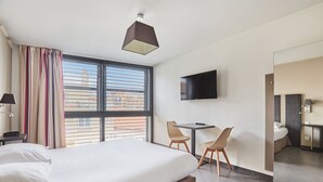 Habitación de hotel con una cama grande, un escritorio con dos sillas, un televisor montado en la pared y una ventana con cortinas.