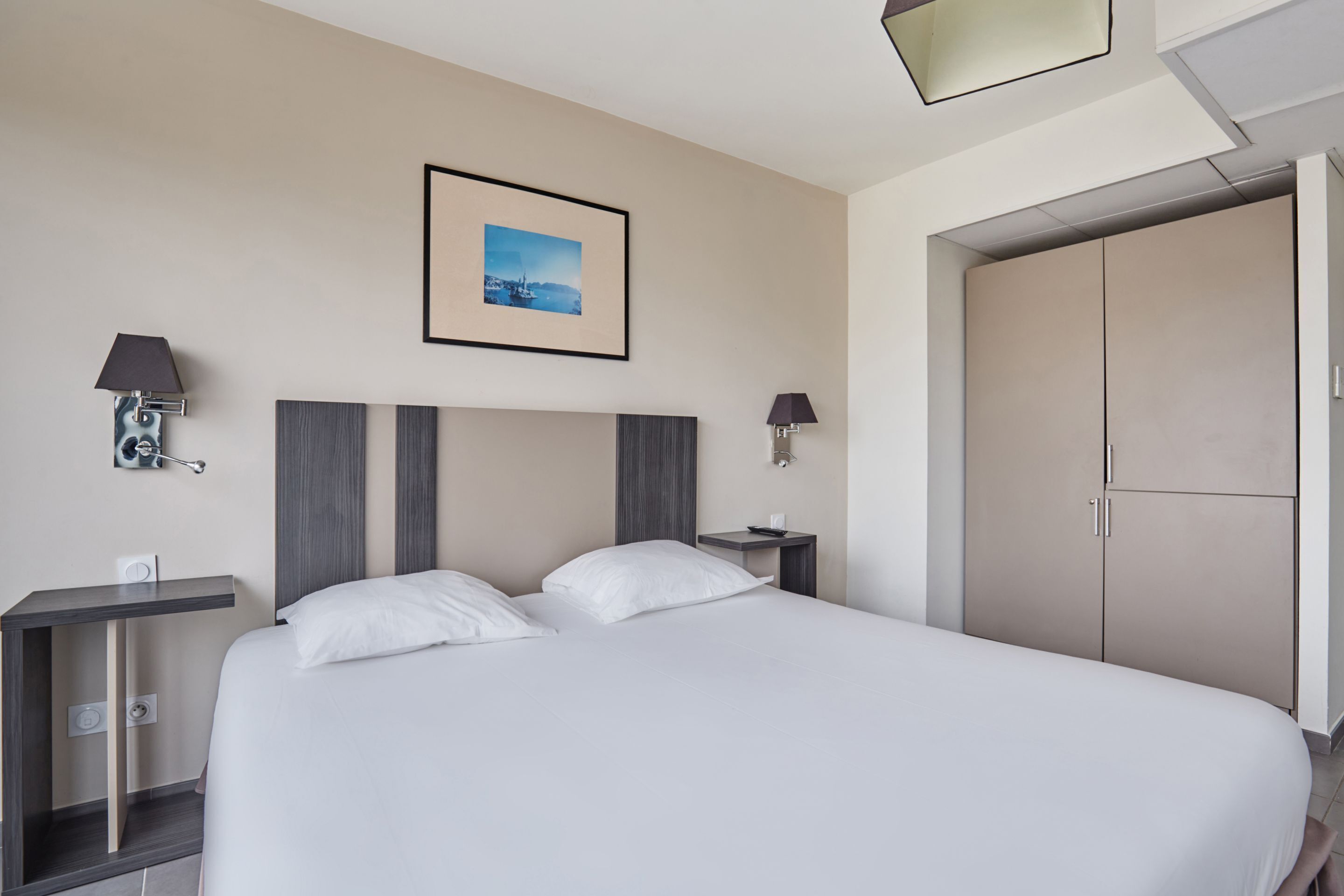 A modern hotel room with a large bed, two bedside tables, a wardrobe, and a framed picture on the wall.