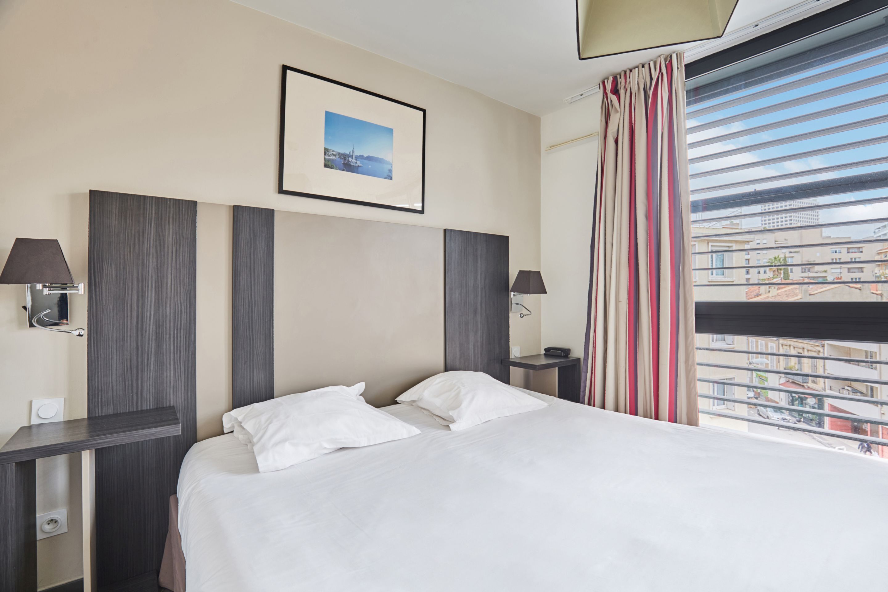 Een hotelkamer met een bed, twee nachtkastjes, een lamp, een fotokader aan de muur en een raam met gordijnen.