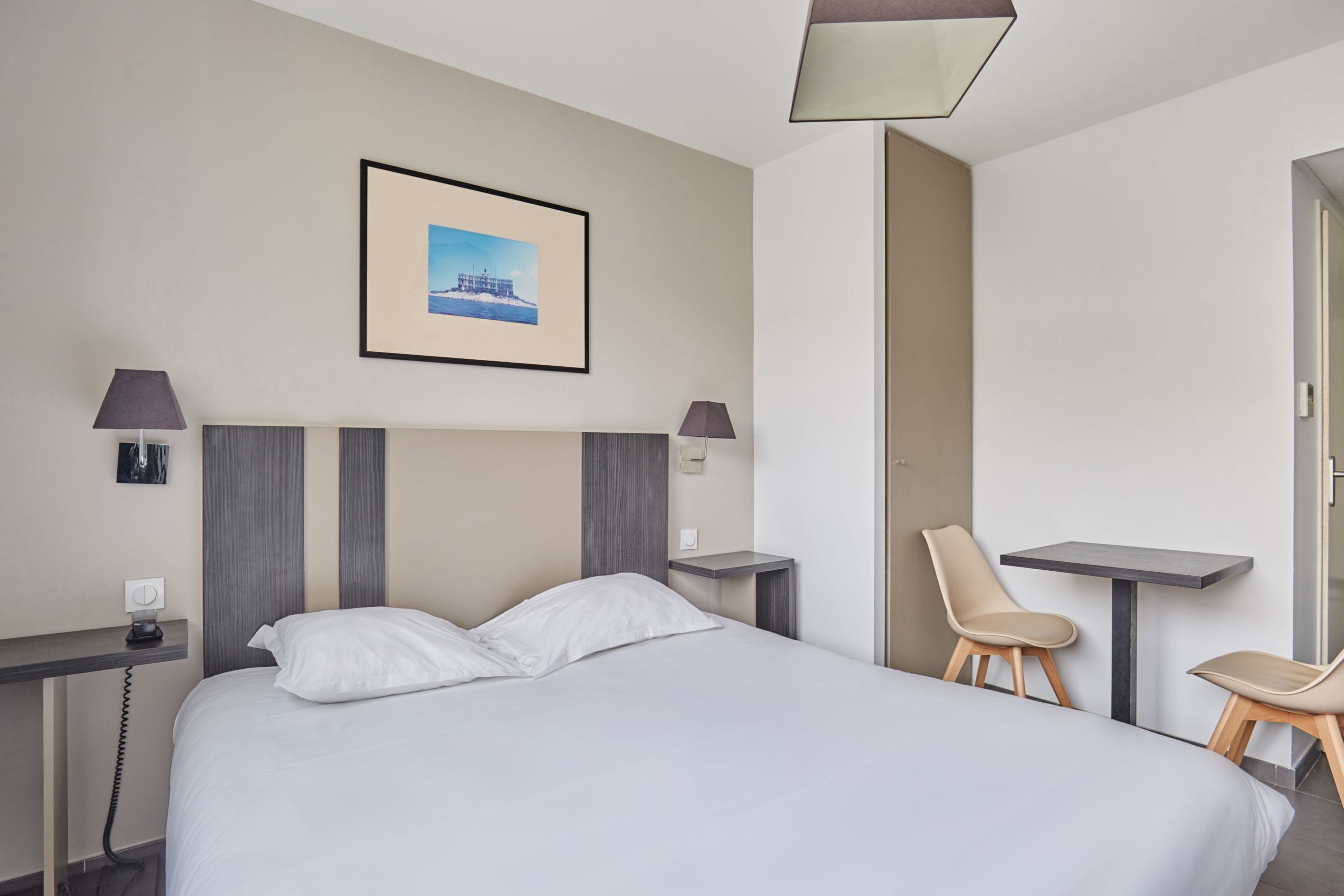 Ein Hotelzimmer mit einem Bett, zwei Sesseln, einem kleinen Tisch, einem Schreibtisch, einer Lampe, einem Bild an der Wand und einem Telefon auf dem Schreibtisch.