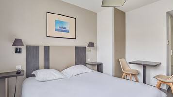 A hotel room with a bed, two chairs, a small table, a desk, a lamp, a picture on the wall, and a phone on the desk.