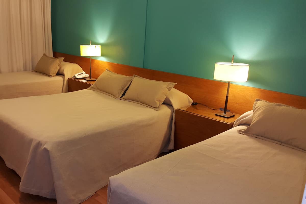 A hotel room with three single beds, each with a bedside table and lamp.