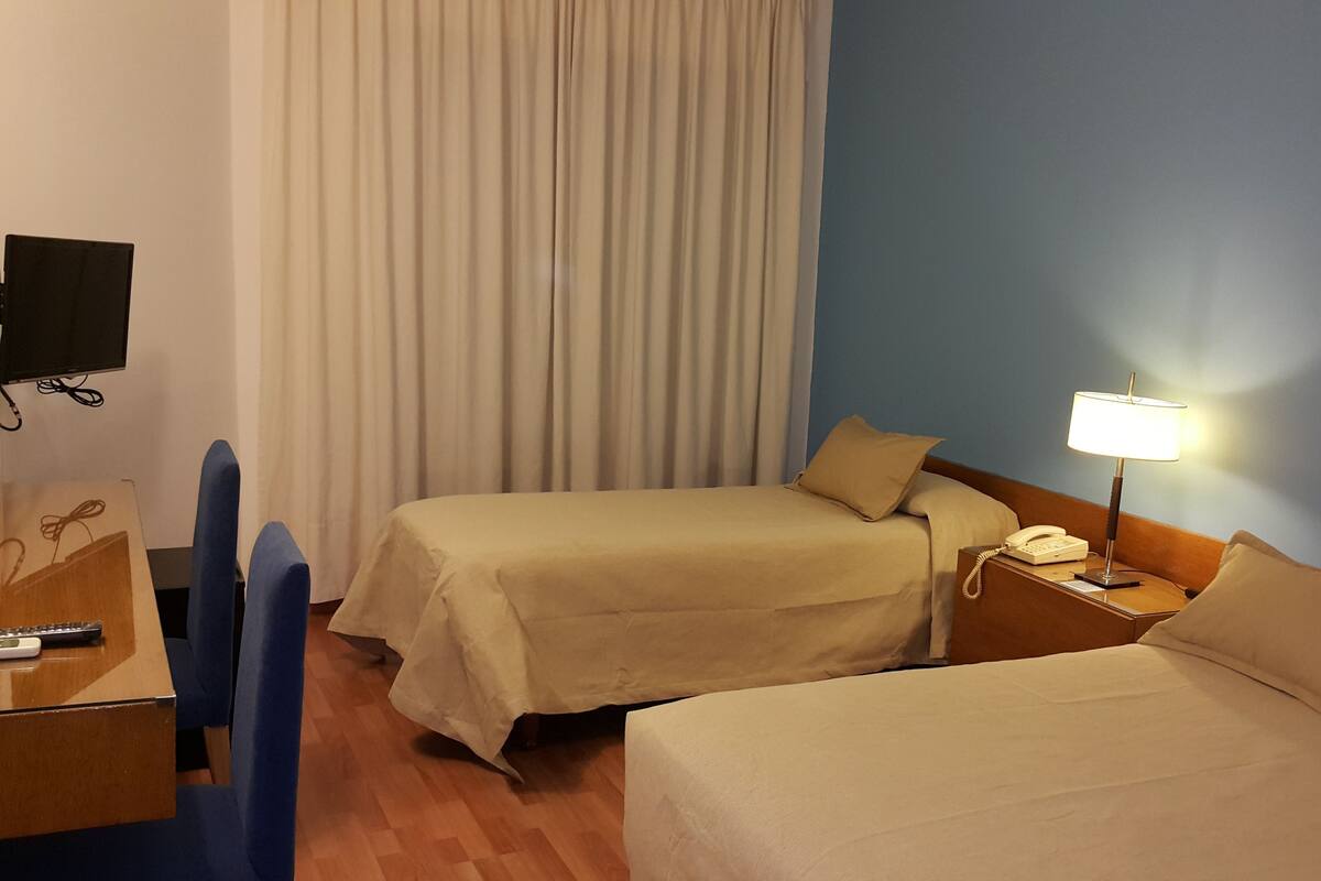 A hotel room with two beds, a desk with a chair, a lamp, and a TV on the wall.