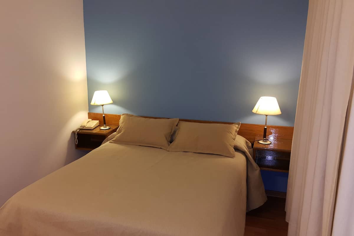 A neatly made bed with a beige bedspread and two pillows, flanked by wooden nightstands with lamps and a telephone.