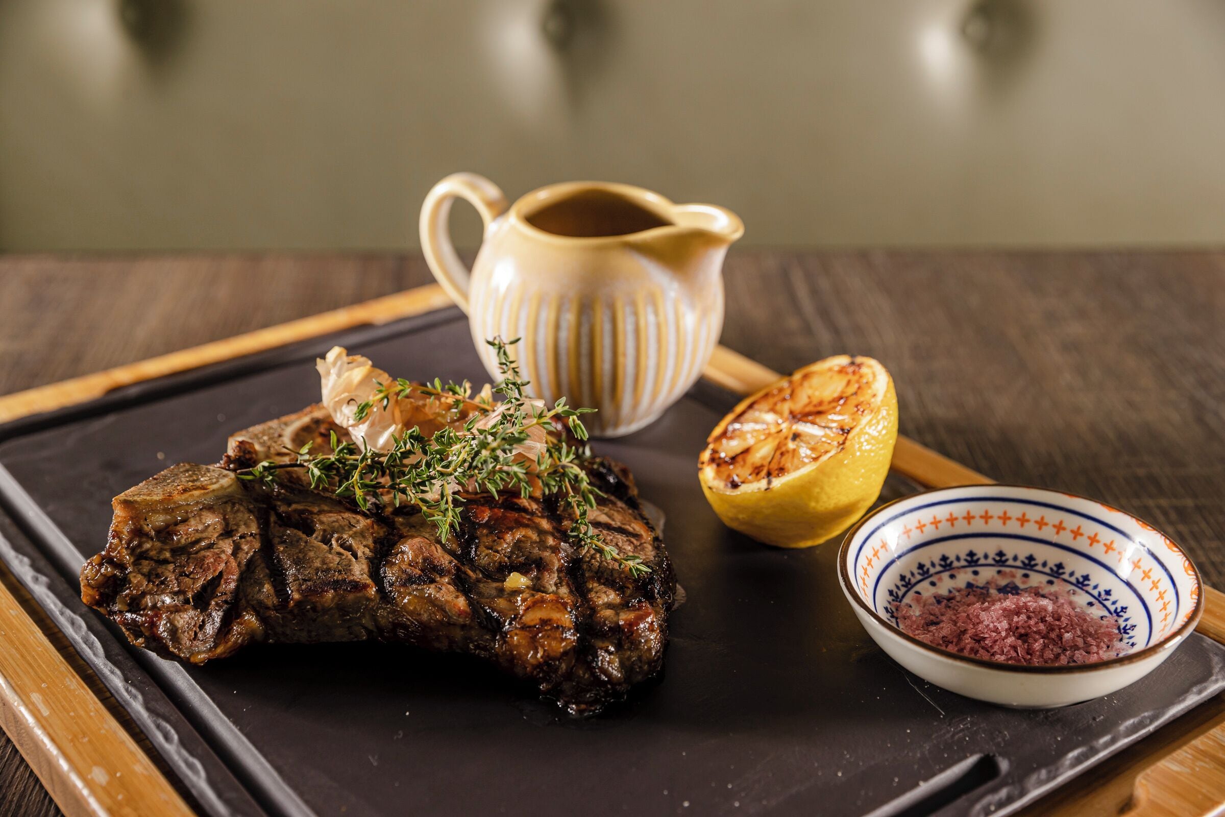 Un bistec con hierbas, media limón, un cuenco de condimentos y una jarras de salsa sobre una bandeja negra.