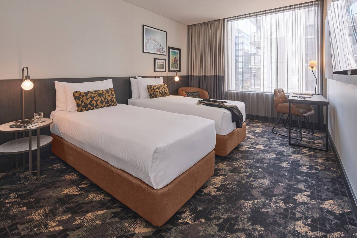 A hotel room with two beds, a small table with a lamp, a chair, and a desk with a chair.