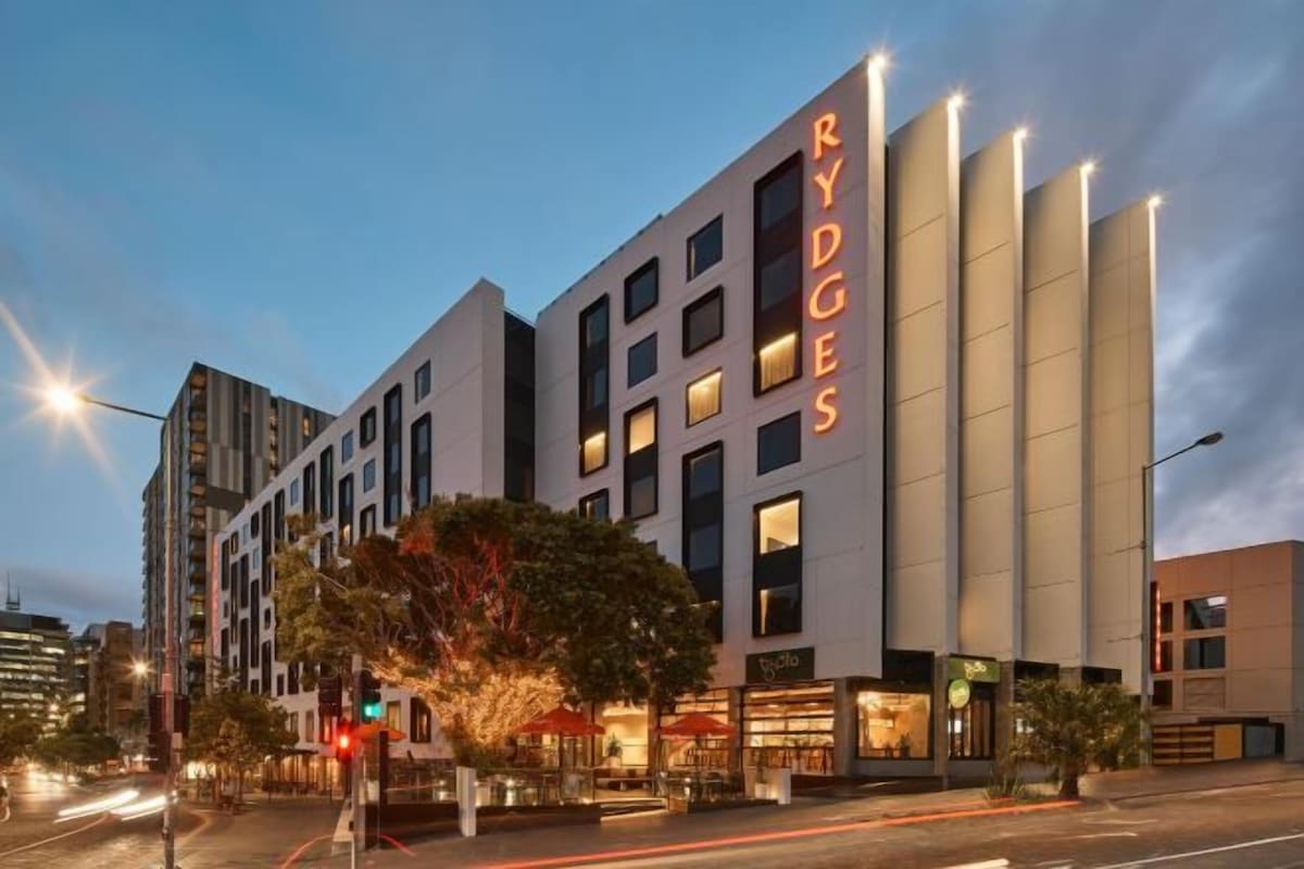 Um hotel moderno com um destaque para a placa 'RYDGES', vários andares e uma área de entrada bem iluminada.