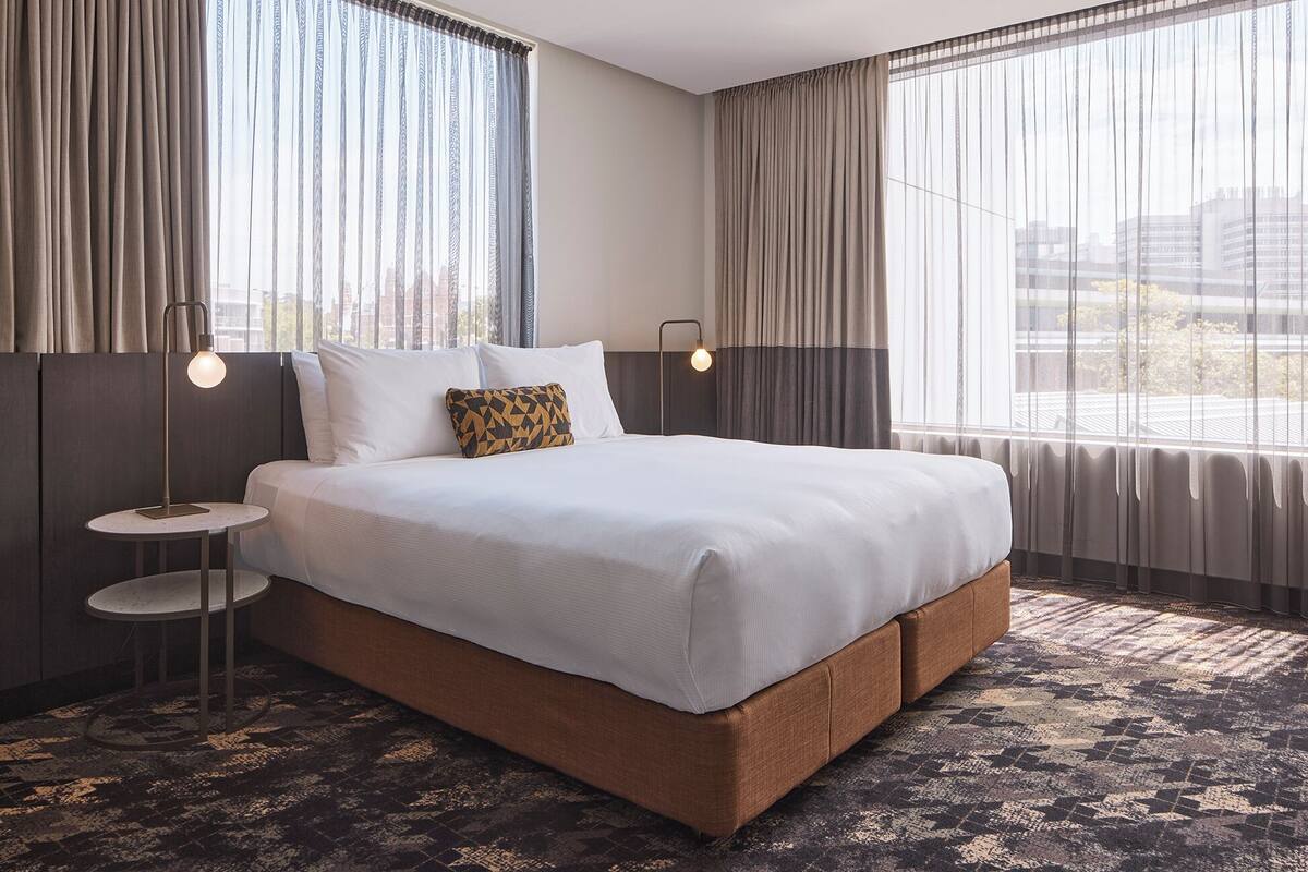 A hotel room with a large bed, two bedside tables, a patterned rug, and a view of buildings through the window.