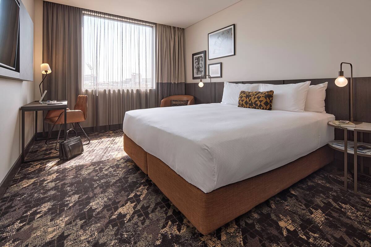 A hotel room with a large bed, a desk with a laptop, a chair, a small table with a lamp, and a window with curtains.
