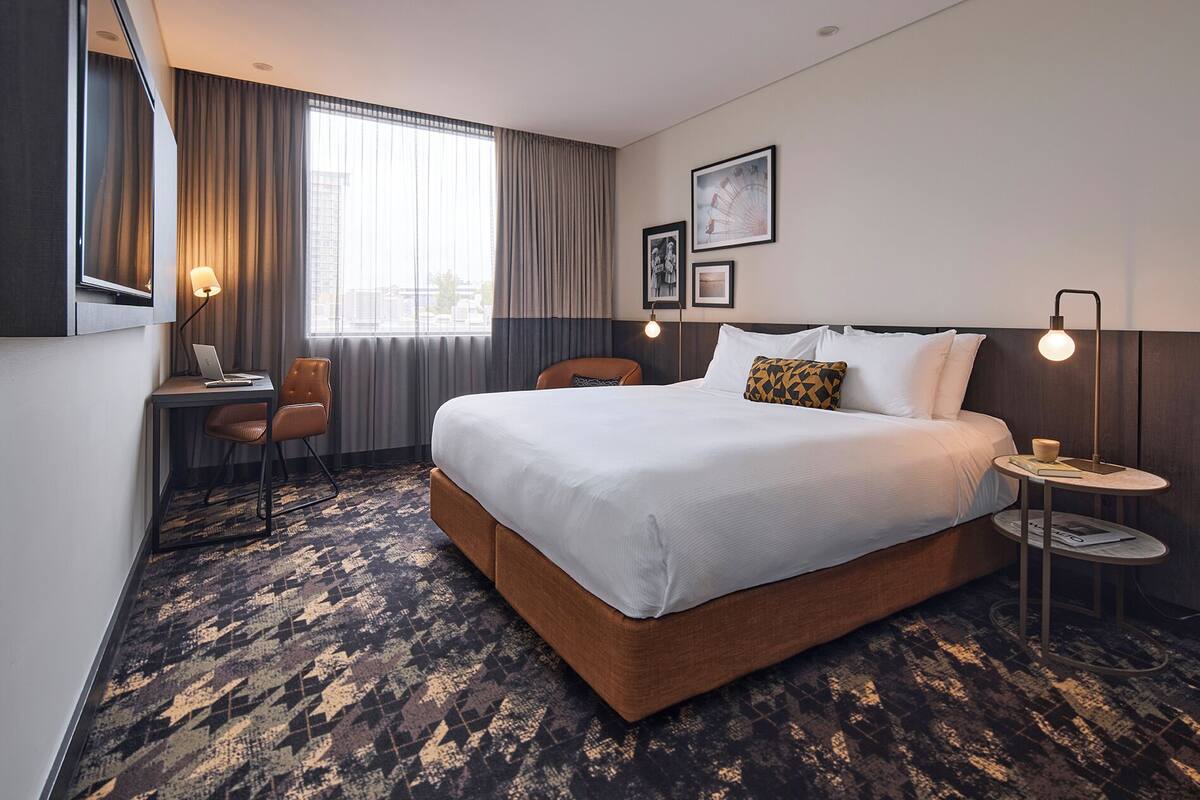 A hotel room with a large bed, a desk, a chair, a small table, and a lamp.