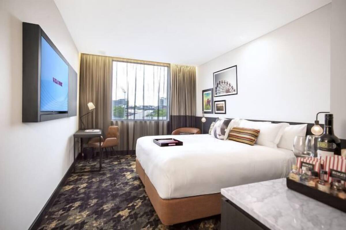 A hotel room with a bed, a desk, a chair, a TV, a window with curtains, and a wall with framed pictures.
