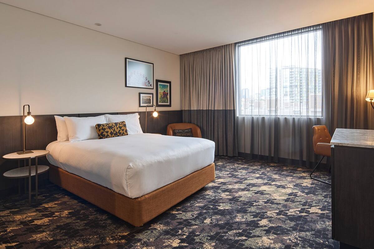 A hotel room with a large bed, a desk, a chair, a round side table, and a view of the city through the window.