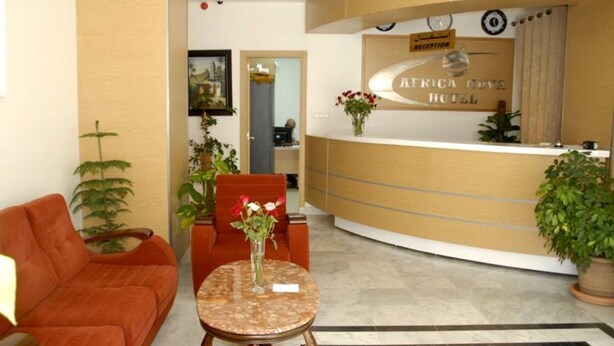 Ein Hotel-Rezeptionsbereich mit einer gebogenen Rezeptionstheke, einem Empfangsschild, einer Couch, einem kleinen runden Tisch mit Blumen und Topfpflanzen.