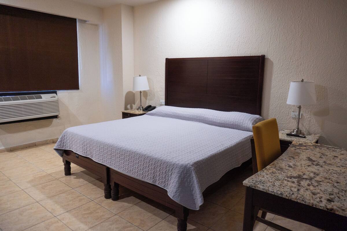 A hotel room with a double bed, a desk with a chair, a lamp, a phone, and a heater.
