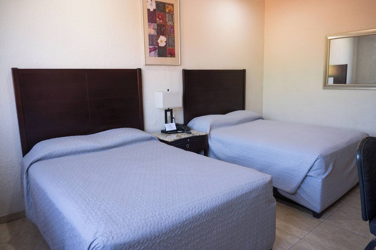 A hotel room with two single beds, a nightstand with a lamp, and a framed picture on the wall.