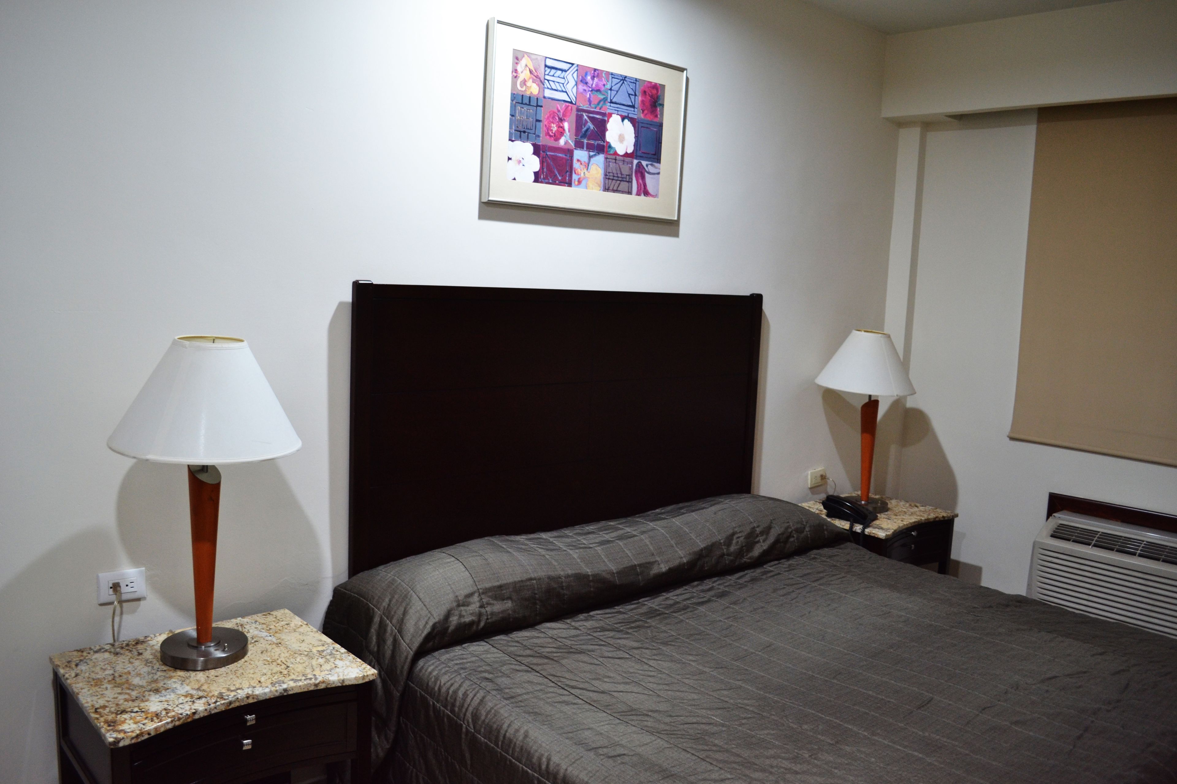 Une chambre d’hôtel comprenant un lit, deux tables de chevet avec des lampes, un tableau accroché au mur et une fenêtre avec des stores.