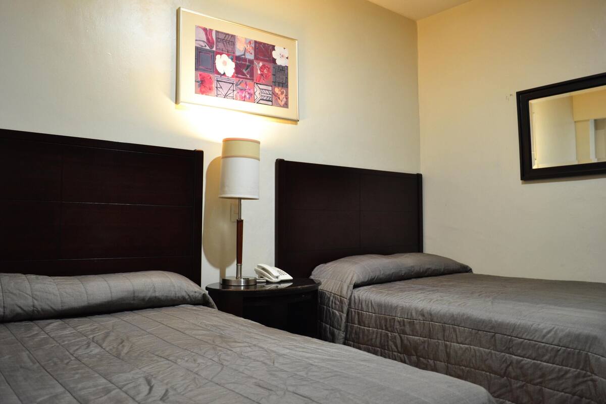 A hotel room with two beds, a bedside table with a lamp, a phone, and a framed picture on the wall.