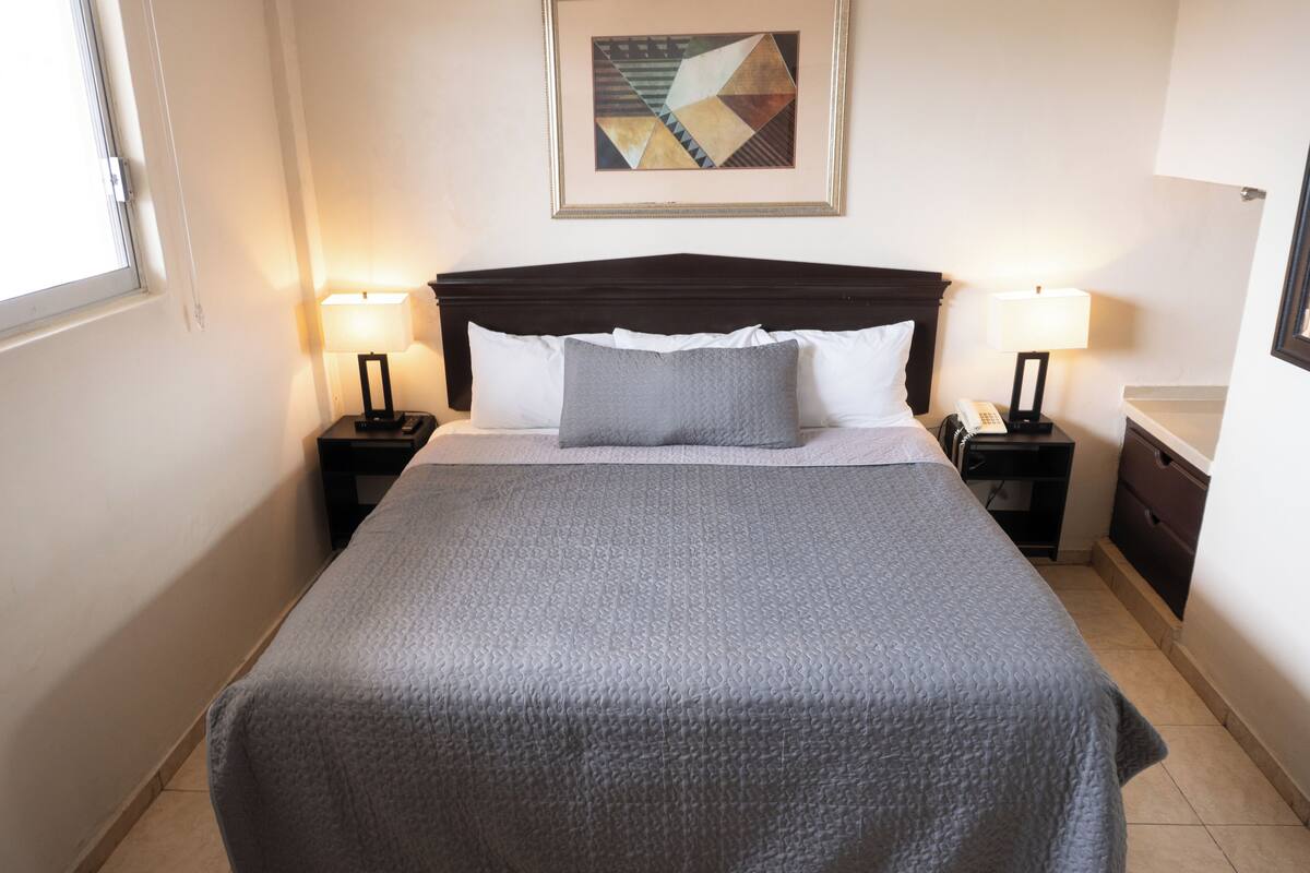 A neatly made bed with a gray quilt, two bedside lamps, a framed abstract painting on the wall, and a small nightstand with a phone.