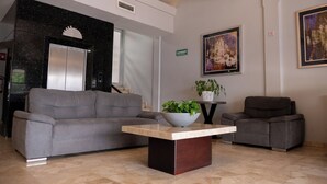 A modern lobby with a grey sofa, a coffee table with a plant, a framed painting, and a digital elevator display. - Hotel America Centro (Los Mochis)