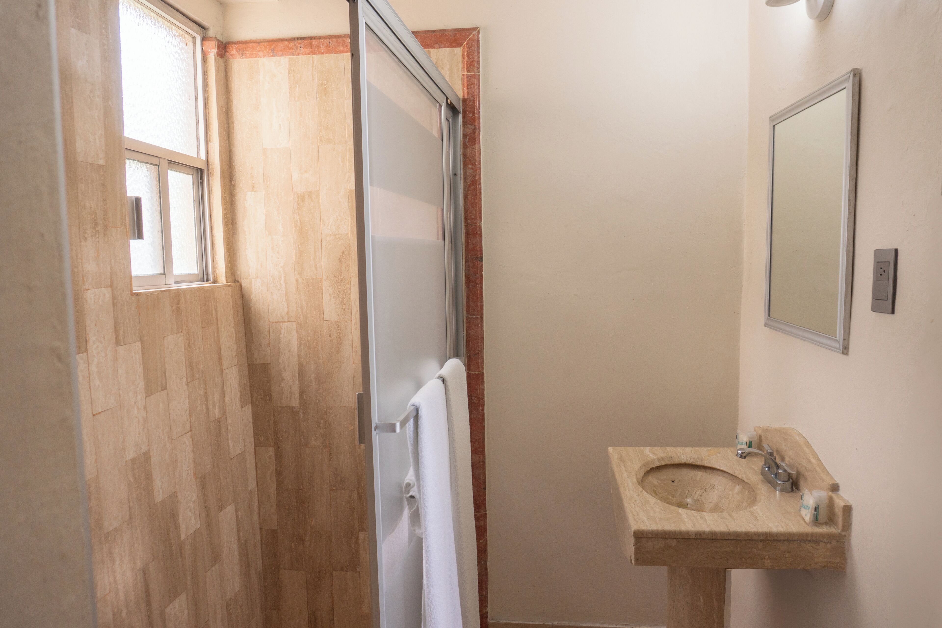 Een badkamer met een houten vloer, een witte handdoek aan een deur, een wastafel met spiegel en een raam.