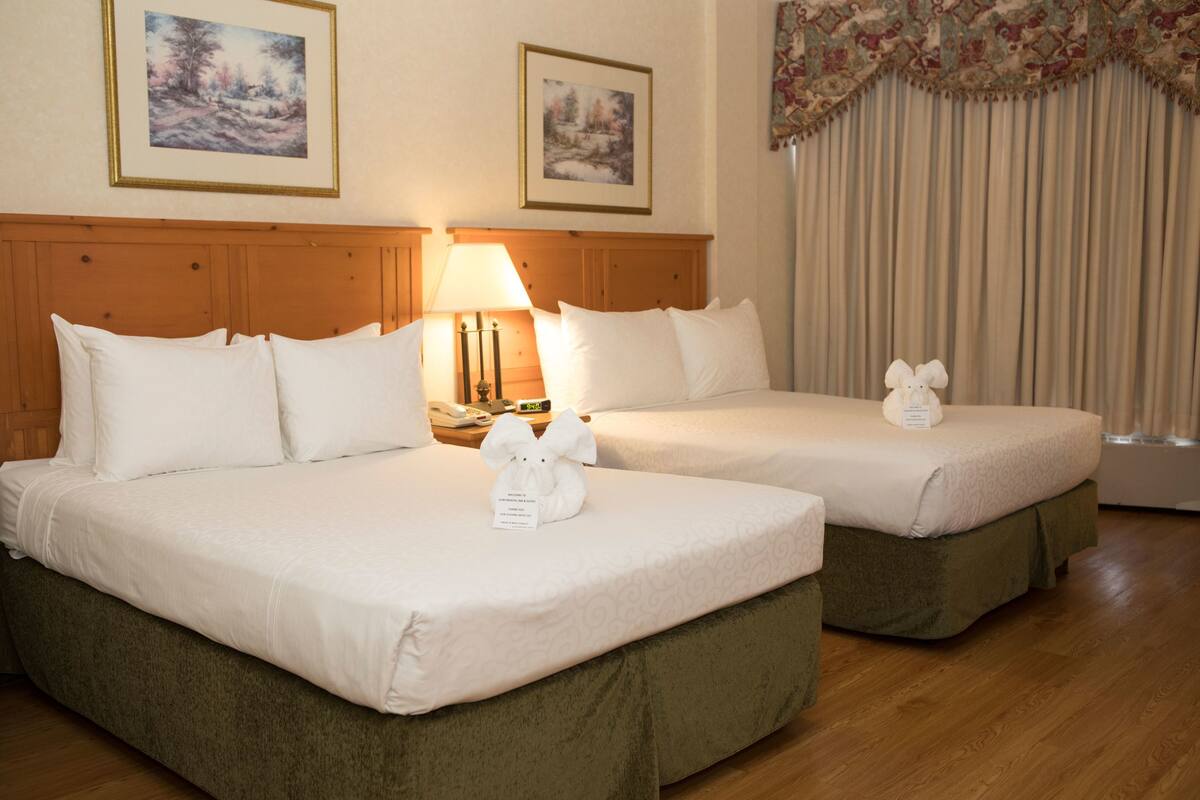 Una habitación de hotel con dos camas, cada una con ropa blanca y falda de cama verde. Hay cuadros en la pared, una lámpara en la mesita de noche y una ventana con cortinas.