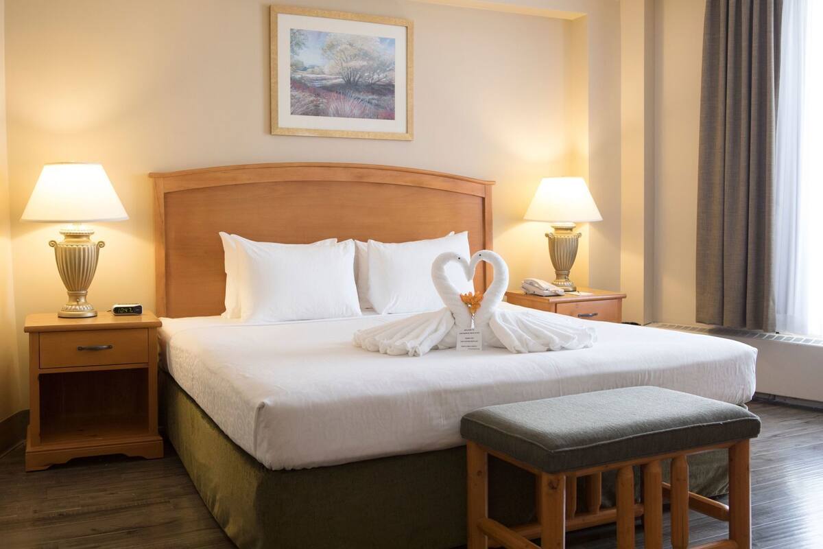Habitación de hotel con una cama grande, dos mesitas de noche con lámparas, un cabecero de madera, un cuadro en la pared y una ventana con cortinas.