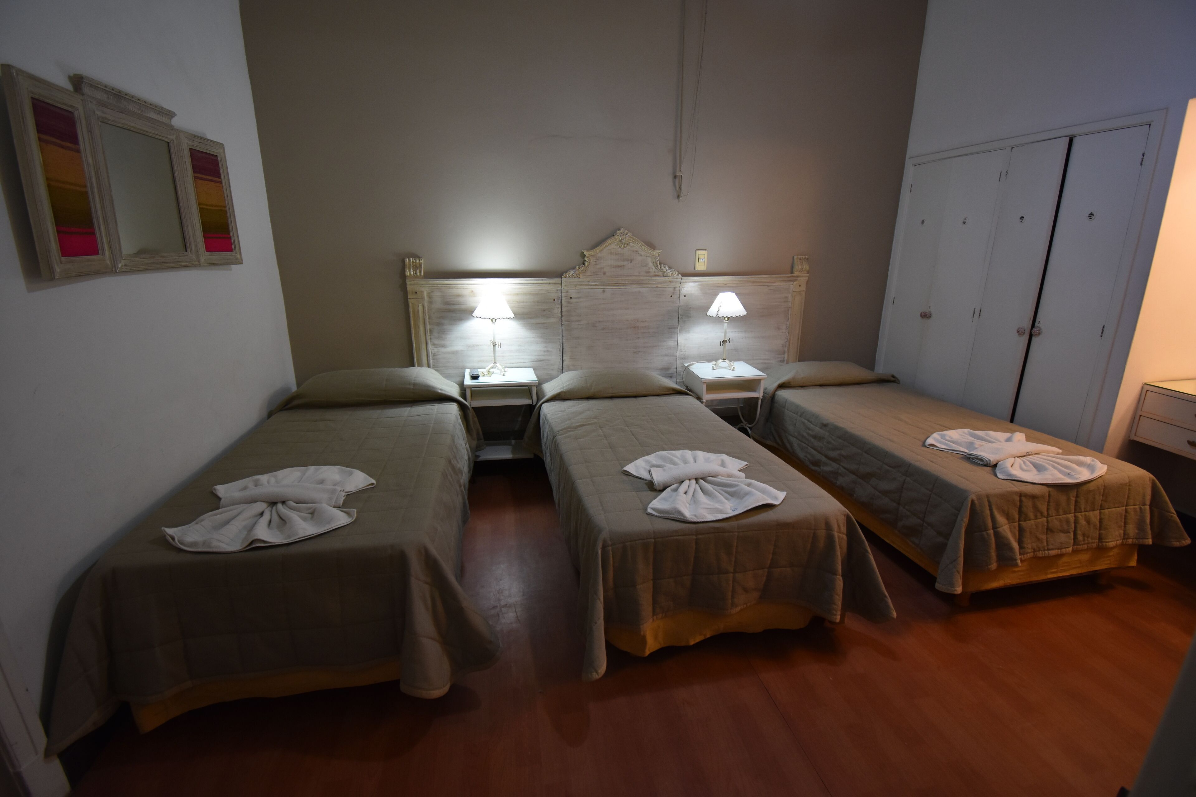 A hotel room with two single beds, each with a pillow and a towel on top. There are bedside tables with lamps, a wardrobe, and a mirror on the wall.