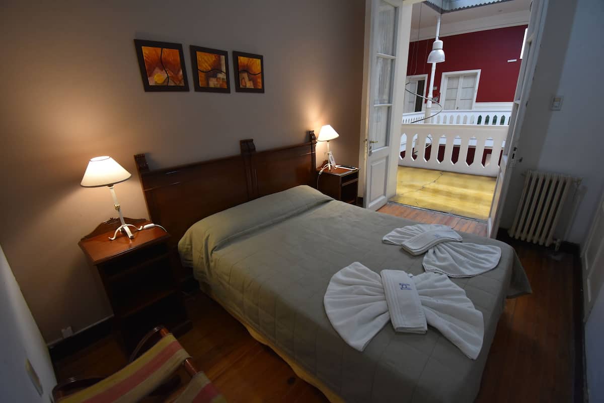 Habitación de hotel con cama, dos lámparas de noche, una mesita de noche de madera, una silla y vistas a un balcón con barandilla blanca.