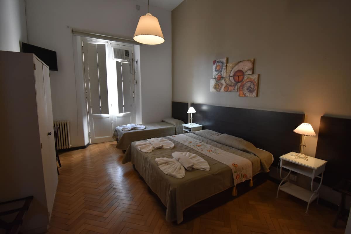 Habitación de hotel con una cama grande, dos mesitas de noche, un armario y una puerta que da a un balcón.