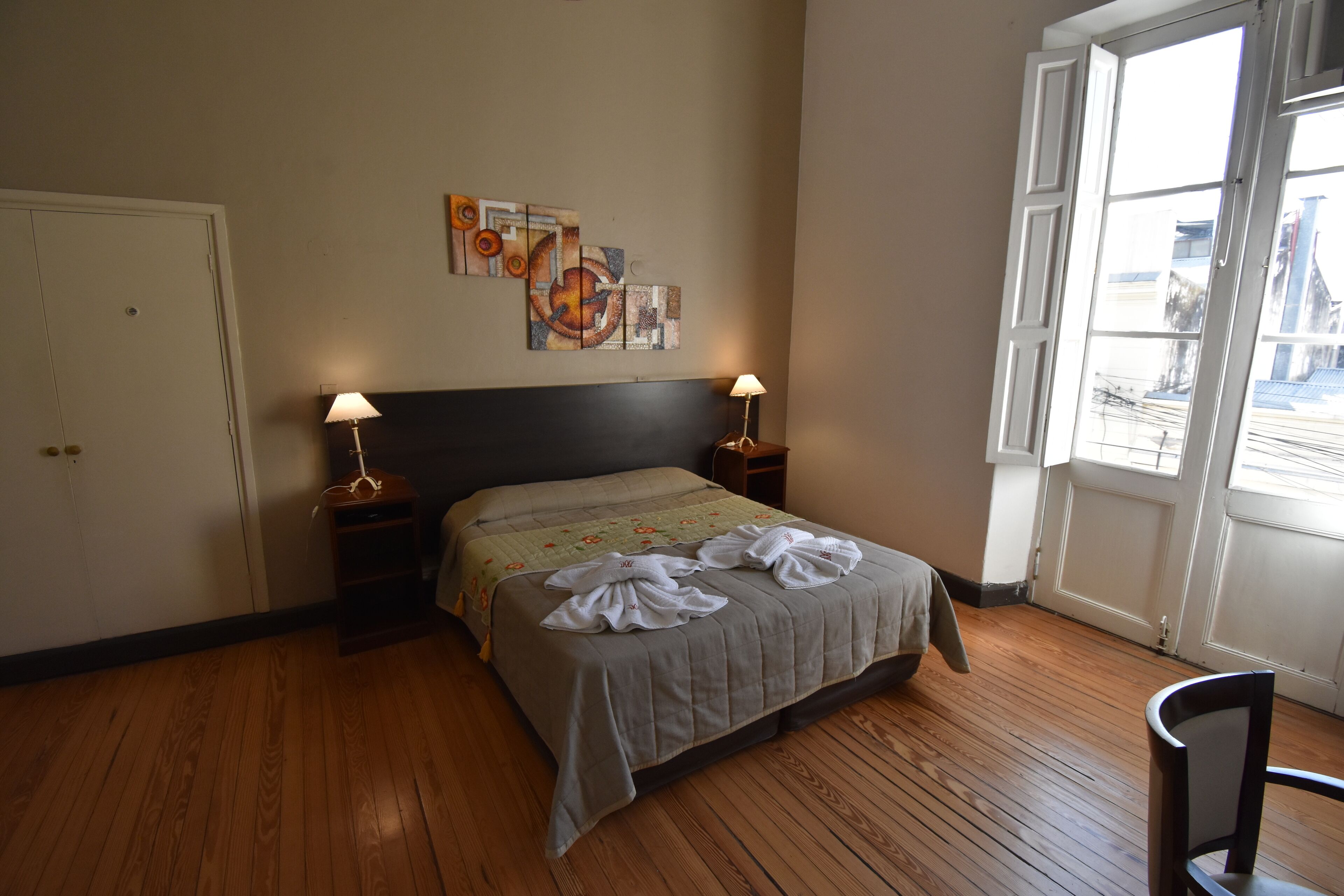 A hotel room with a wooden floor, a bed with a grey bedspread, two bedside lamps, a wall art piece, and a window with a view of buildings.
