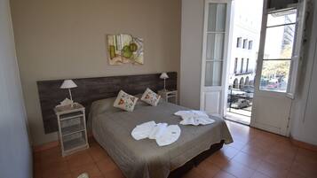 Quarto com cama, travesseiros, mesa de cabeceira, luminária e janela com vista para edifícios e rua.