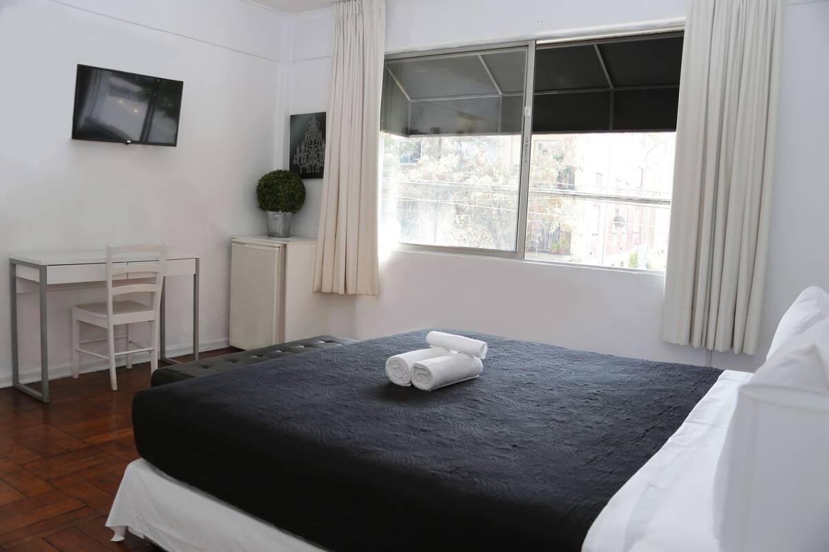 Habitación de hotel con cama, escritorio, silla, televisor y ventana con cortinas.