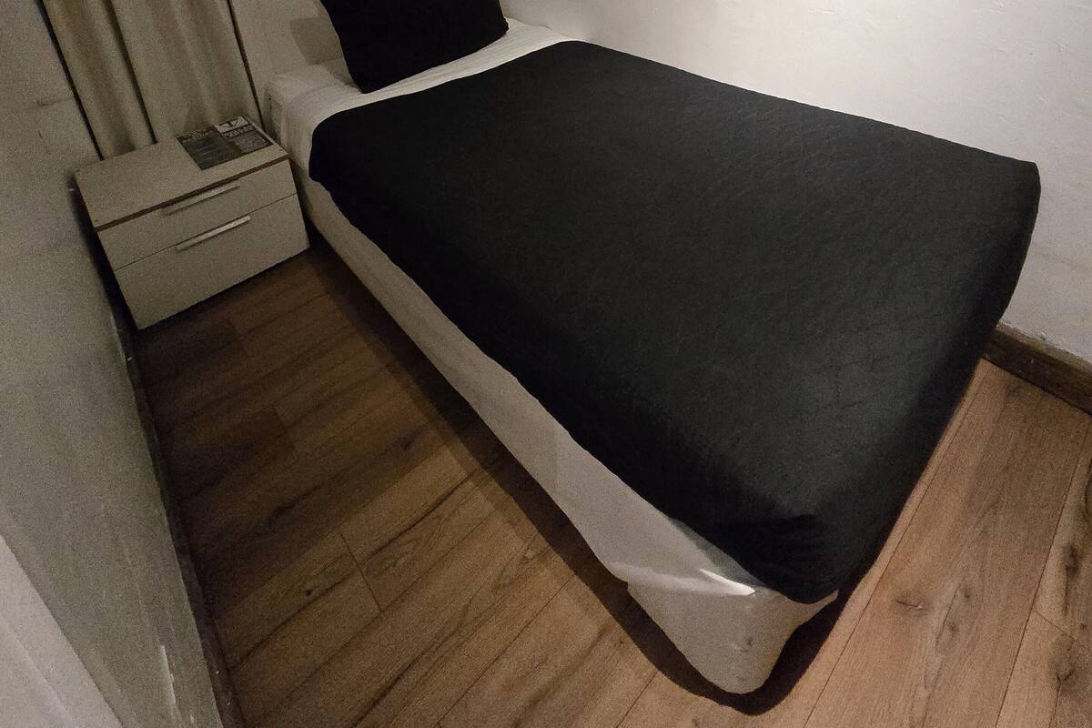 Una habitación pequeña con cama individual, suelo de madera, una mesita de noche blanca y una ventana con cortinas.