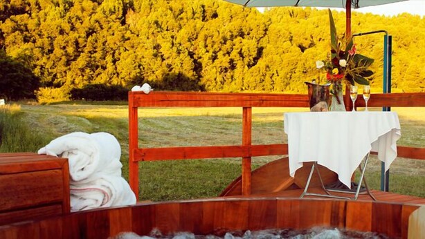 Tinaco de madera con un mantel blanco y un florero, sobre un fondo de follaje amarillo.