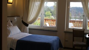 Un dormitorio con cama, dos ventanas con cortinas, una silla y vistas a una terraza y árboles.