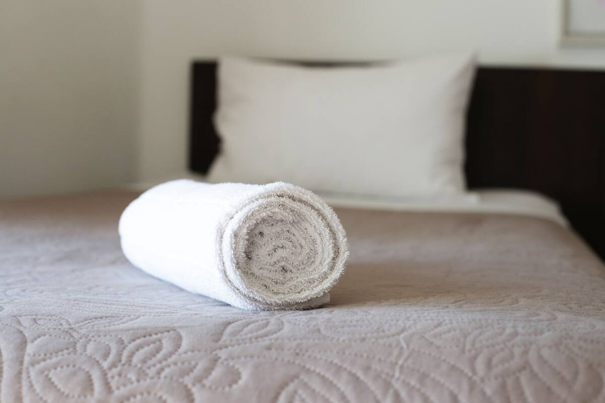 Un lit bien fait, avec une serviette blanche roulée posée sur le dessus, un oreiller blanc et une tête de lit sombre.