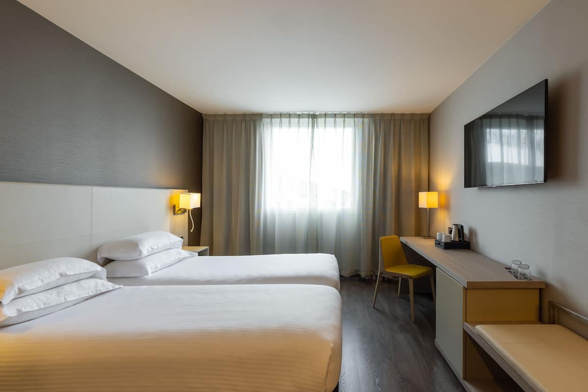A hotel room with two beds, a desk, a chair, a TV, and a window with curtains.