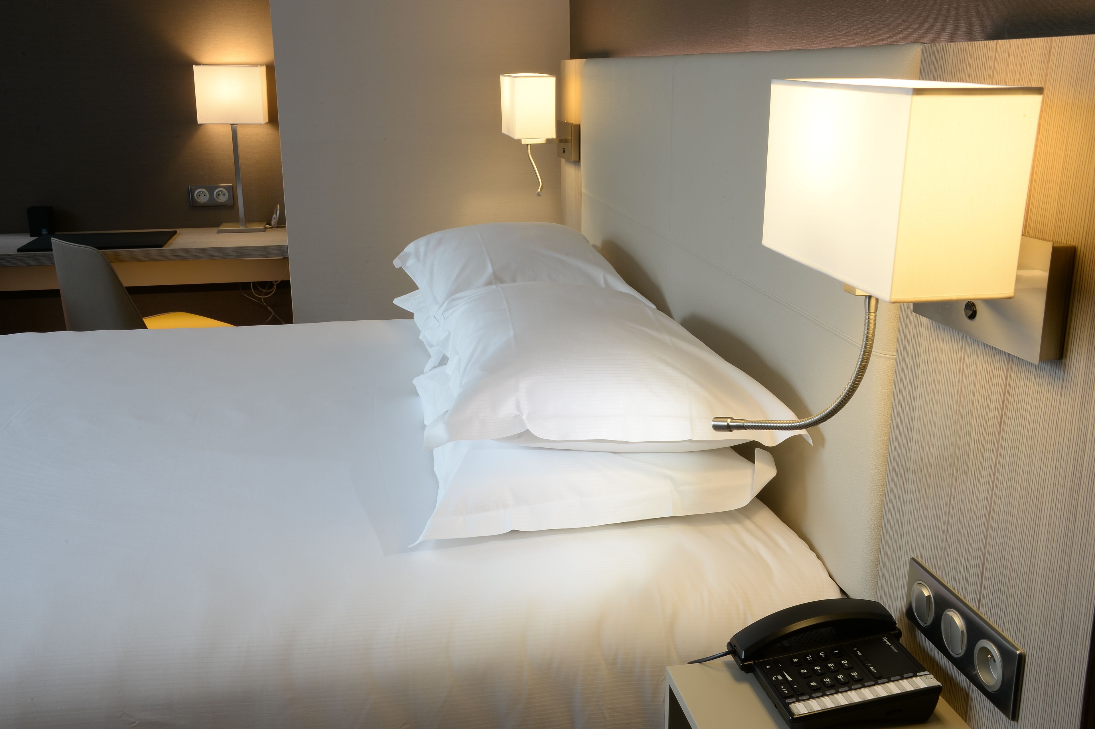 Een netjes opgemaakt bed met witte lakens, een bedlampje, een telefoon en een klein bureau met een lamp en een klok.