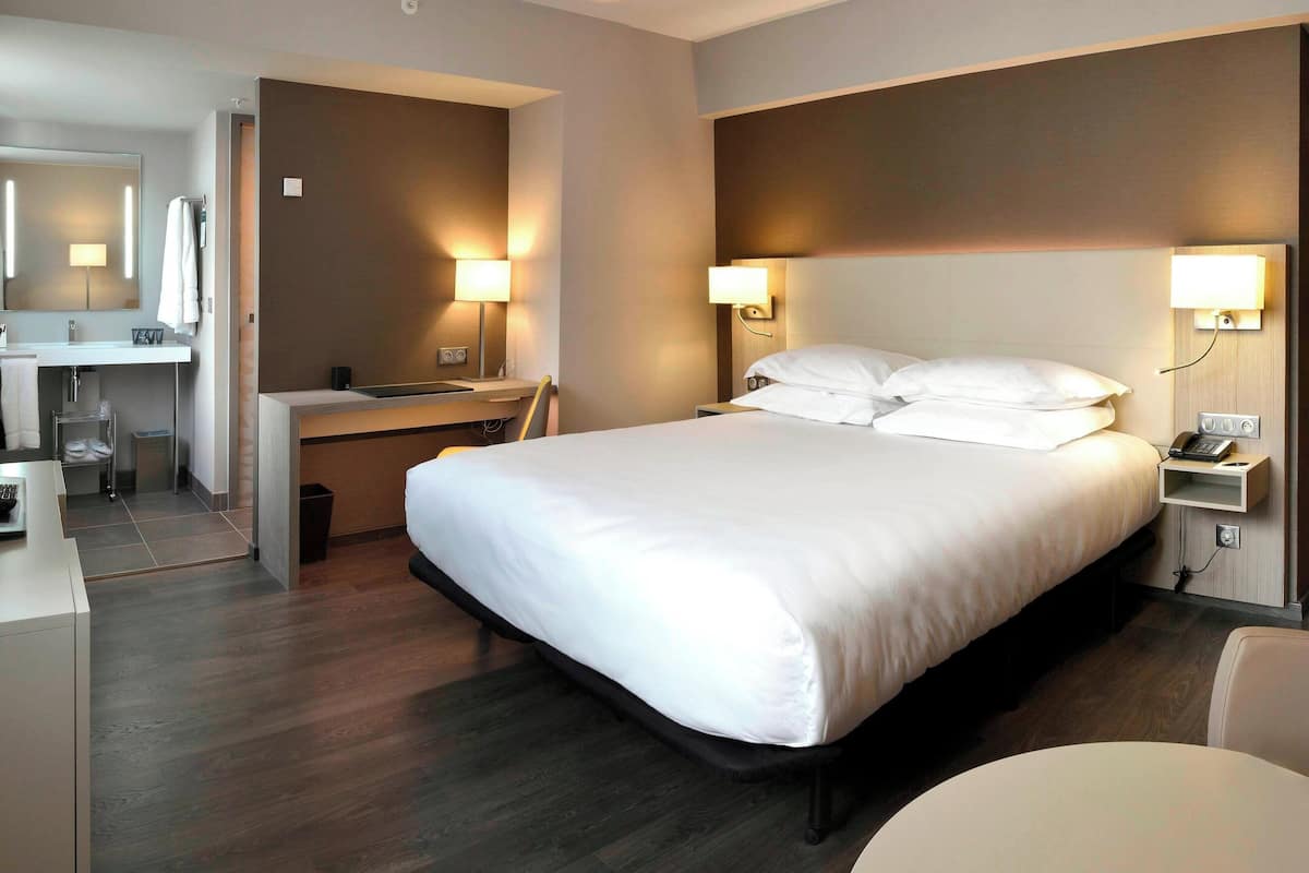 A modern hotel room with a large bed, a desk with a chair, a vanity with a mirror, and bedside lamps.