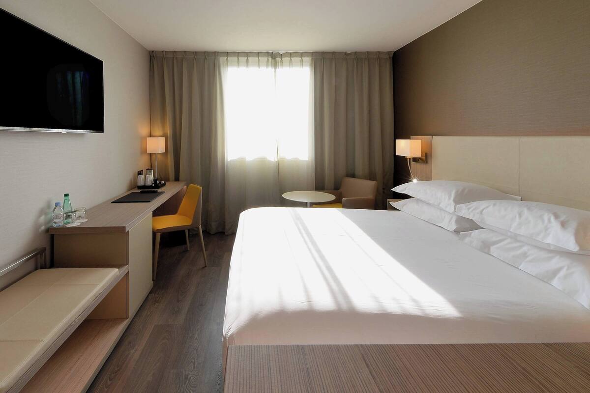 A hotel room with a large bed, a desk, a chair, a small table, and a TV mounted on the wall.