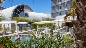 A poolside area with lounge chairs and umbrellas, a modern building with a large circular window, and a palm tree in the foreground. - AC Hotel by Marriott Marseille Prado Velodrome (Marseille)