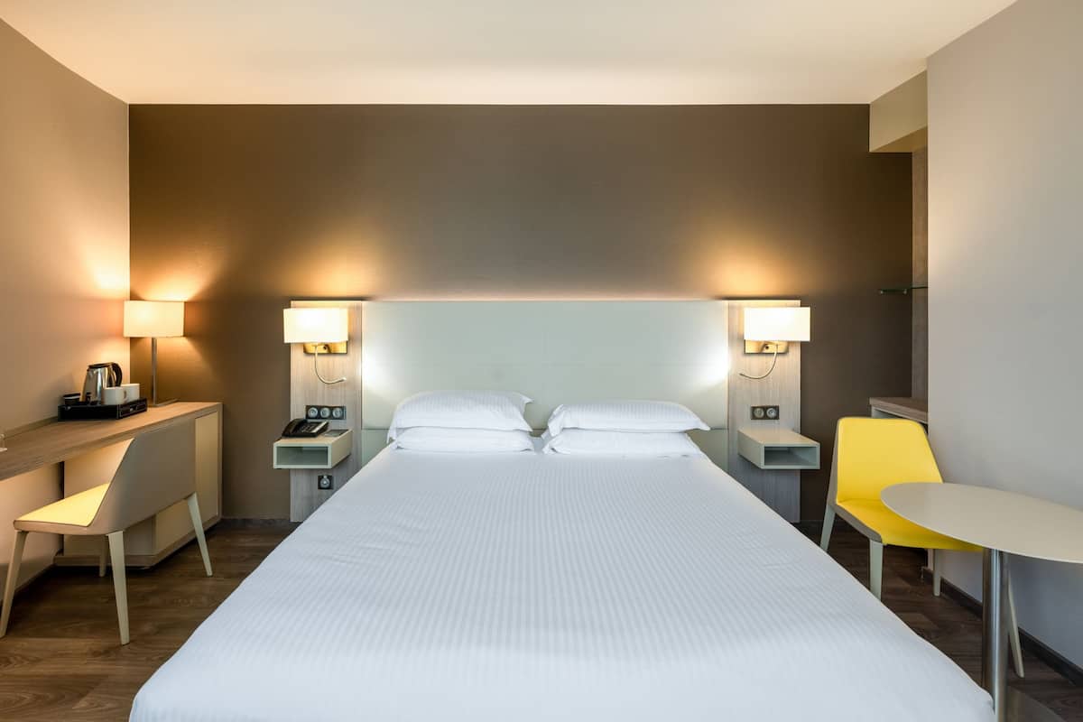 A hotel room with a large bed, bedside lamps, a desk with a chair, and a small table with a chair.