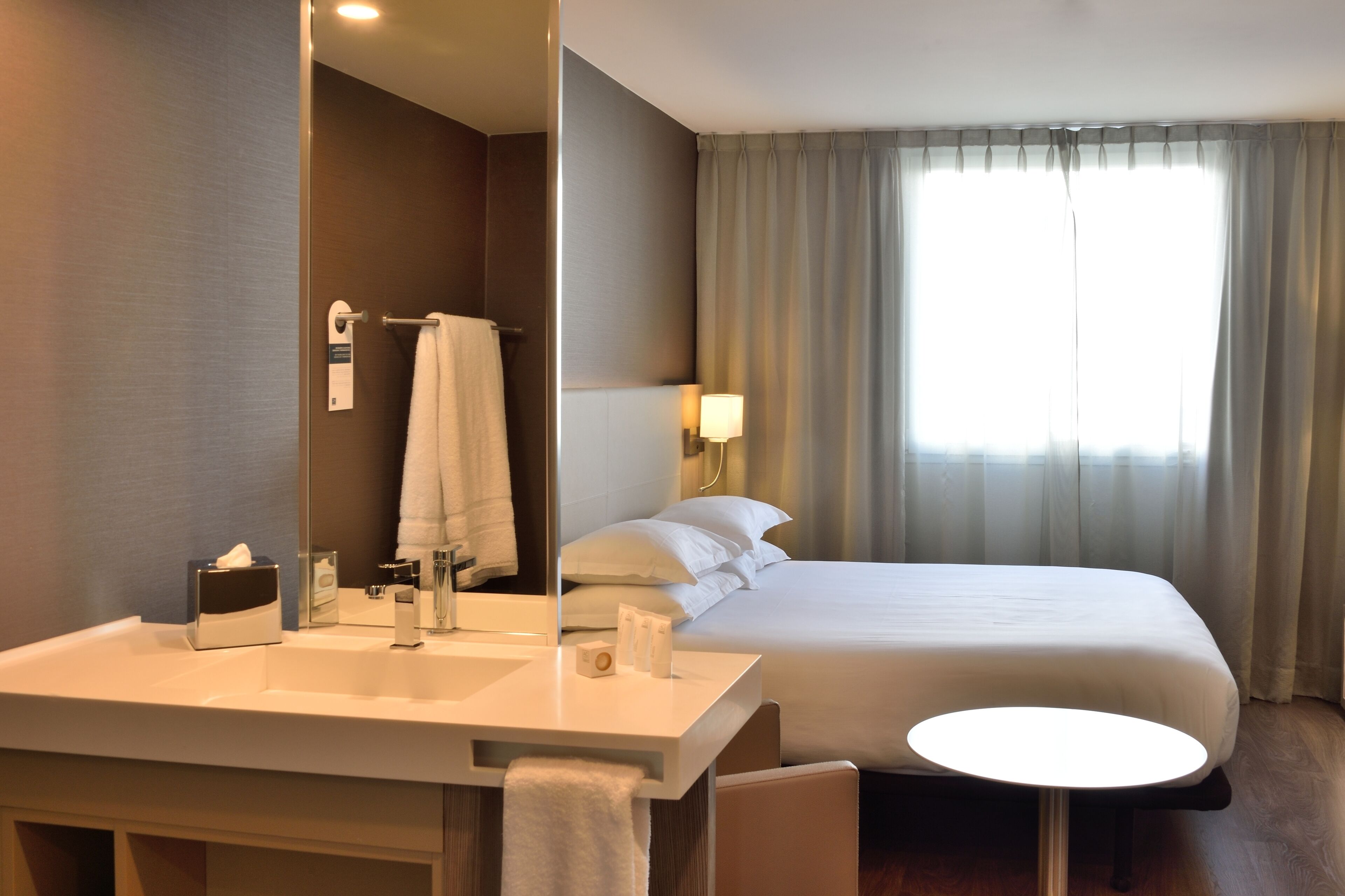 A hotel room with a large bed, a round table, a bathroom with a sink and mirror, and a window with curtains.